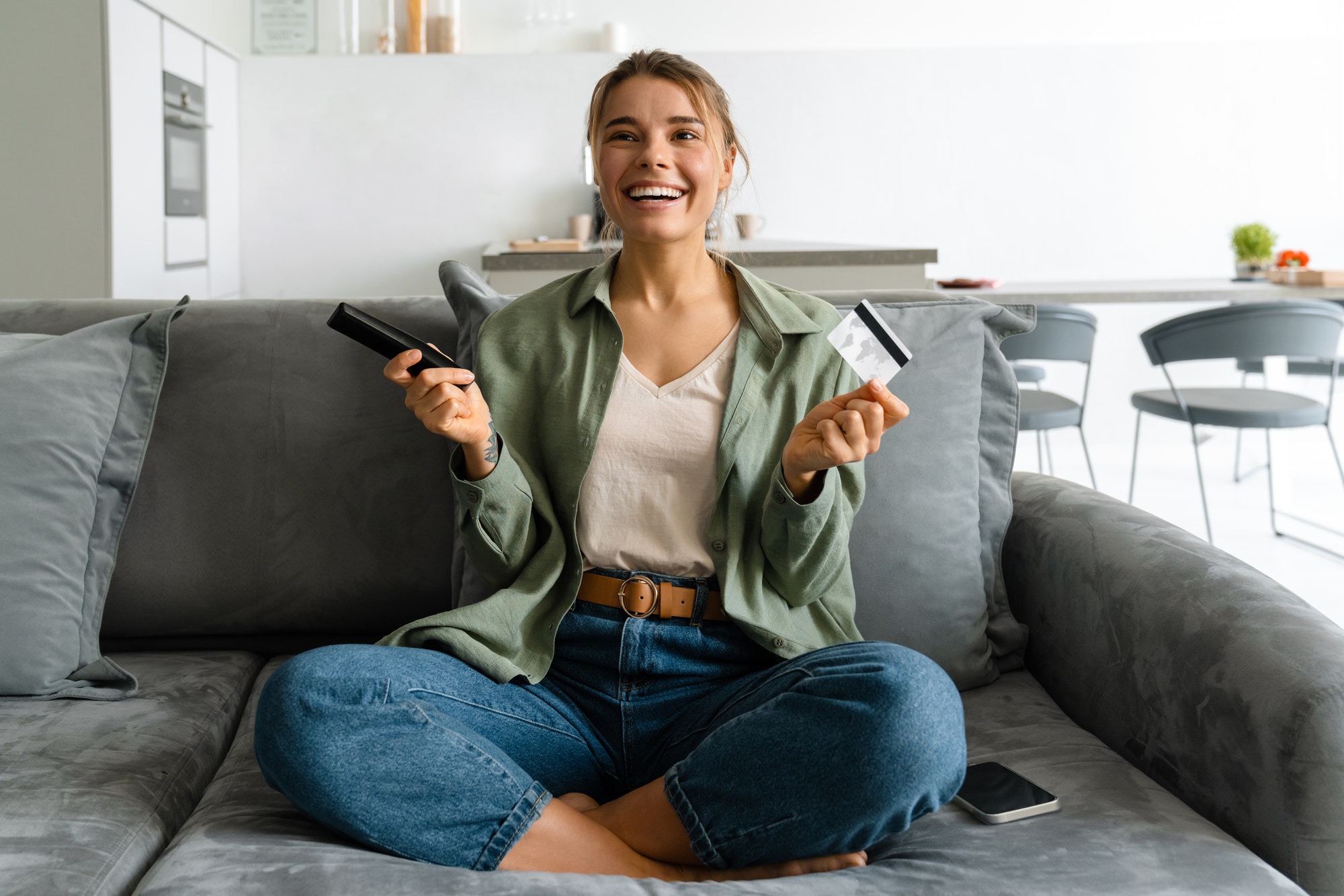 Happy woman holding credit card while watching TV with remote control